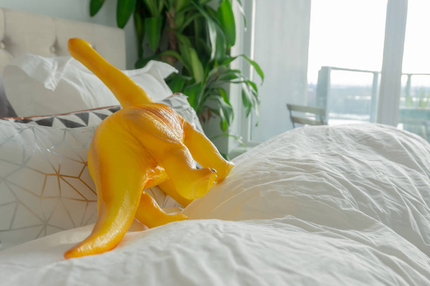 Gelber Spielzeug-Dinosaurier im Elternbett, der den Lebensstil der Eltern und ein richtiges Zuhause darstellt. - Foto, Bild