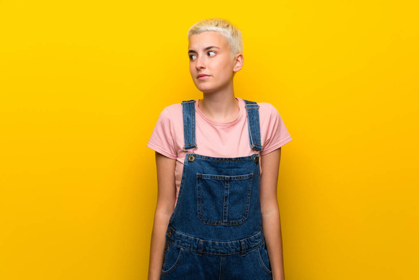 Teenager girl with overalls on yellow background making doubts gesture looking side - Foto, afbeelding