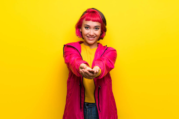 Jovem mulher com cabelo rosa sobre a parede amarela segurando copyspace imaginário na palma da mão para inserir um anúncio
 - Foto, Imagem