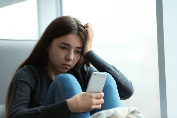 Upset teenage girl with smartphone sitting at window indoors. Space for text - Foto, afbeelding