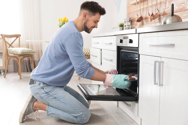 Handsome man taking out tray of baked cookies from oven in kitchen - Photo, image