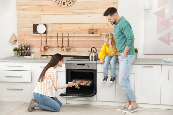 Woman and her family taking out tray with baked cookies from oven in kitchen - Zdjęcie, obraz