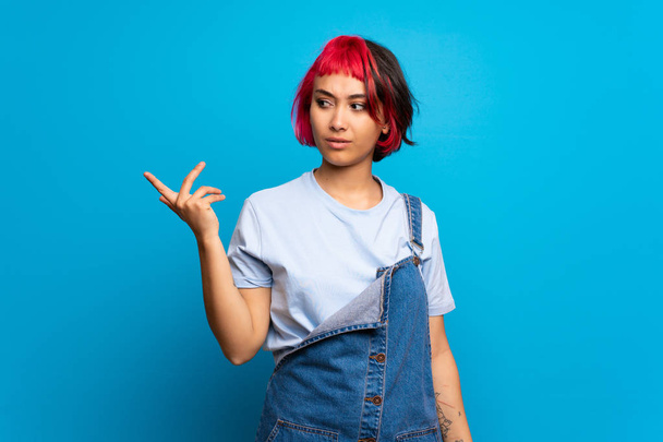 Молодая женщина с розовыми волосами над синей стеной делает жесты сомнений
 - Фото, изображение
