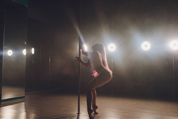 Young slim woman pole dancing in dark interior with lights and smoke. - Фото, изображение