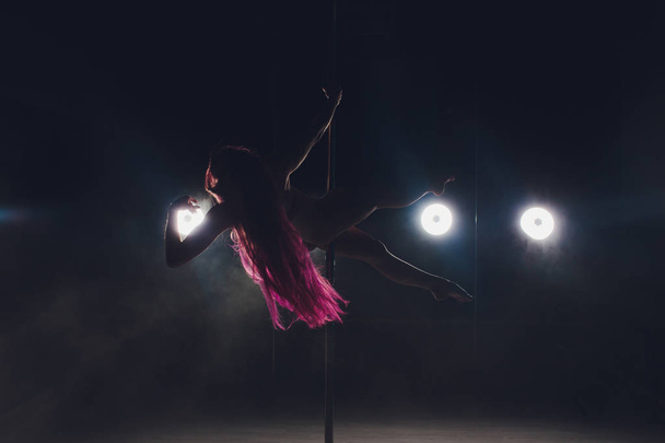 Νεαρή λεπτή γυναίκα πόλο χορεύει σε σκοτεινό εσωτερικό με φώτα και καπνό. - Φωτογραφία, εικόνα