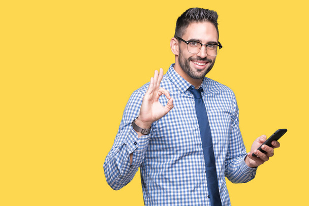 Young business man using smartphone over isolated background doing ok sign with fingers, excellent symbol - Foto, imagen