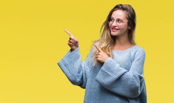 Young beautiful blonde woman wearing sweater and glasses over isolated background smiling and looking at the camera pointing with two hands and fingers to the side. - 写真・画像