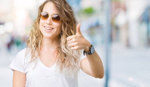 Beautiful young blonde woman wearing sunglasses over isolated background doing happy thumbs up gesture with hand. Approving expression looking at the camera with showing success. - Photo, Image
