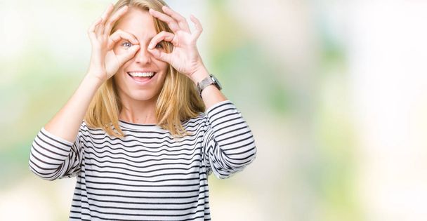 Beautiful young woman wearing stripes sweater over isolated background doing ok gesture like binoculars sticking tongue out, eyes looking through fingers. Crazy expression. - Foto, Bild