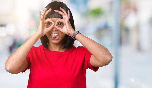 Krásná mladá africká americká žena nosí brýle izolované pozadí dělá ok gesto jako dalekohled držet jazyk, oči dívá skrze prsty. Šílený výraz. - Fotografie, Obrázek