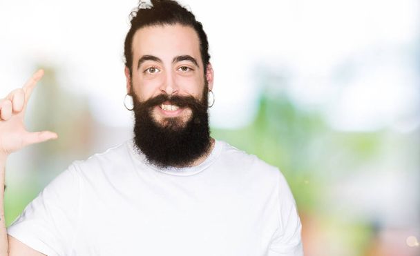 Young hipster man with long hair and beard wearing casual white t-shirt smiling and confident gesturing with hand doing size sign with fingers while looking and the camera. Measure concept. - Фото, изображение