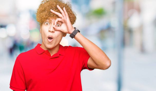 Young handsome man with afro hair wearing red t-shirt doing ok gesture shocked with surprised face, eye looking through fingers. Unbelieving expression. - Foto, afbeelding