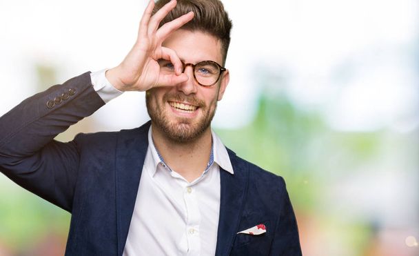 Young handsome business man wearing glasses doing ok gesture with hand smiling, eye looking through fingers with happy face. - 写真・画像