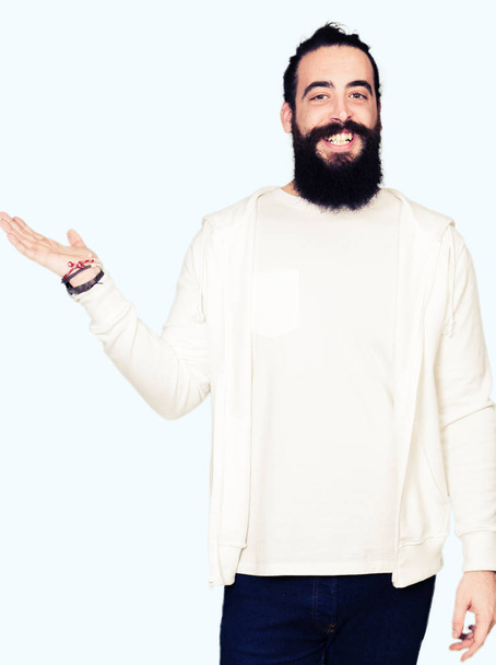 junger Mann mit langen Haaren und Bart, der ein sportliches Sweatshirt trägt, fröhlich lächelt und mit der Handfläche in die Kamera zeigt. - Foto, Bild