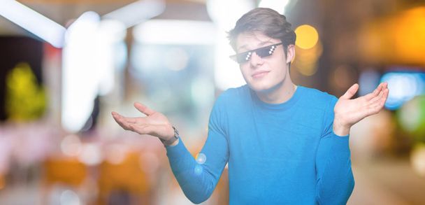 Young man wearing funny thug life glasses over isolated background clueless and confused expression with arms and hands raised. Doubt concept. - Photo, Image