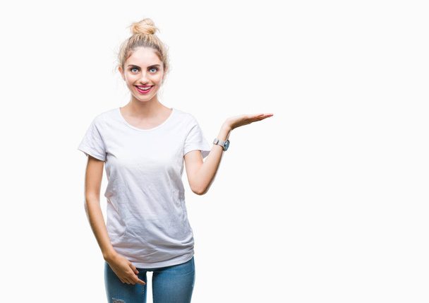 Young beautiful blonde woman wearing white t-shirt over isolated background smiling cheerful presenting and pointing with palm of hand looking at the camera. - Fotoğraf, Görsel