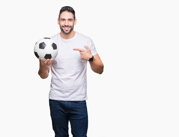 Jonge man houdt van voetbal voetbal over geïsoleerde achtergrond erg blij met de hand en de vinger te wijzen - Foto, afbeelding