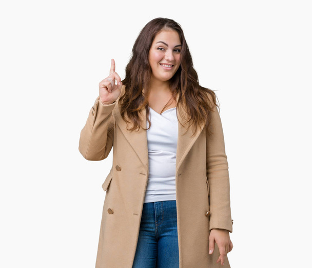 Mooie plus grootte jonge vrouw winterjas te dragen over geïsoleerd achtergrond wijzende vinger omhoog met succesvolle idee. Verlaten en gelukkig. Nummer één. - Foto, afbeelding