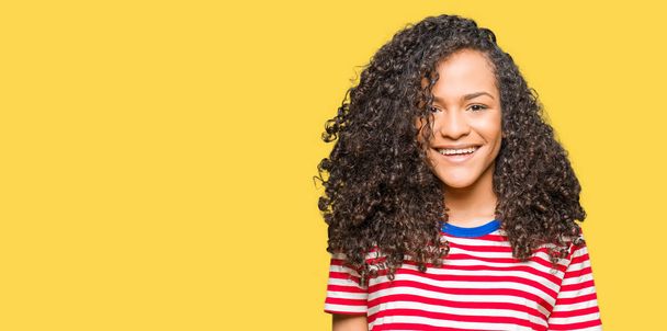Young beautiful woman with curly hair wearing stripes t-shirt with a happy and cool smile on face. Lucky person. - Photo, Image