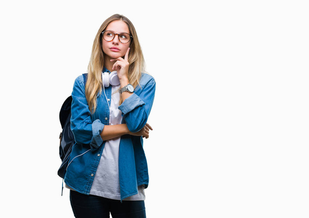 Young beautiful blonde student woman wearing headphones and glasses over isolated background with hand on chin thinking about question, pensive expression. Smiling with thoughtful face. Doubt concept. - Photo, image
