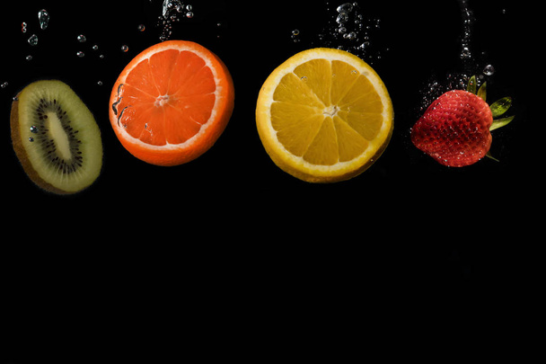 mehrere Früchte hintereinander: Kiwi, Orange, Zitrone und Erdbeere, in Wasser getaucht mit Blasen auf schwarzem Hintergrund. - Foto, Bild