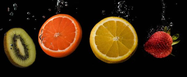 Kilka owoców z rzędu: kiwi, pomarańcza, cytryna i truskawka, zanurzone w wodzie z bąbelkami na czarnym tle. - Zdjęcie, obraz
