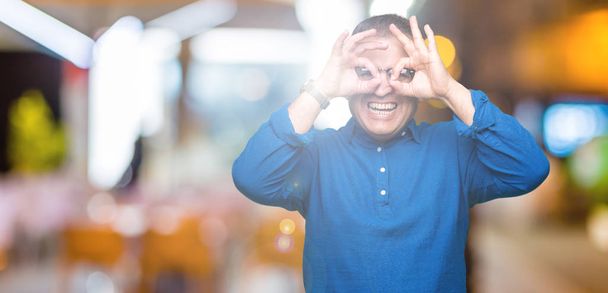 Hombre árabe de mediana edad con gafas sobre un fondo aislado haciendo un buen gesto como prismáticos sacando la lengua, ojos mirando a través de los dedos. Expresión loca
. - Foto, Imagen