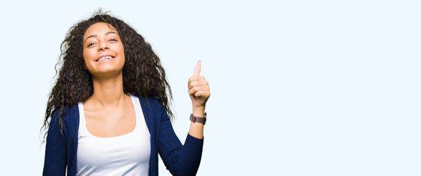 Young beautiful girl with curly hair doing happy thumbs up gesture with hand. Approving expression looking at the camera with showing success. - Foto, Imagen