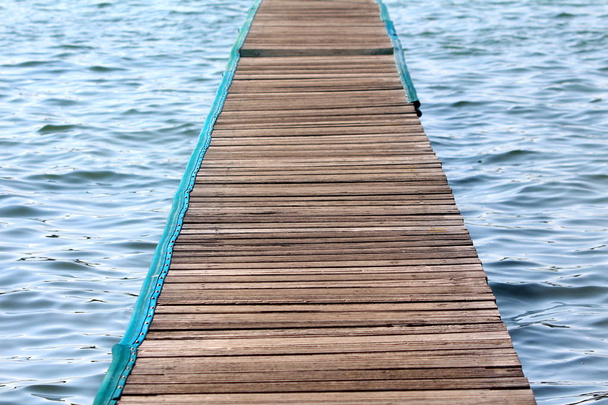 Деревянный пирс из нескольких узких досок с нейлоновой защитой по краям над спокойным голубым морем в теплый солнечный день
 - Фото, изображение