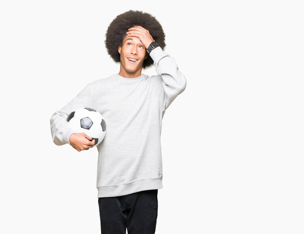 Νεαρός αφρικανική αμερικανική άνθρωπος με τα άφρο μαλλιά, κρατώντας την μπάλα ποδοσφαίρου ποδοσφαίρου τόνισε με το χέρι στο κεφάλι, σοκαρισμένος με ντροπή και έκπληξη πρόσωπο, θυμωμένος και απογοητευμένος. Φόβο και αναστάτωση για το λάθος. - Φωτογραφία, εικόνα