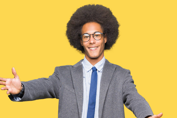 Jeune homme d'affaires afro-américain aux cheveux afro portant des lunettes regardant la caméra souriant à bras ouverts pour un câlin. Expression joyeuse embrassant le bonheur
. - Photo, image