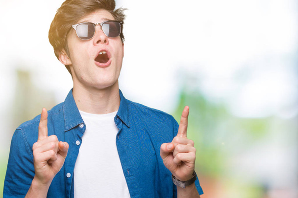 Young handsome man wearing sunglasses over isolated background amazed and surprised looking up and pointing with fingers and raised arms. - Photo, Image