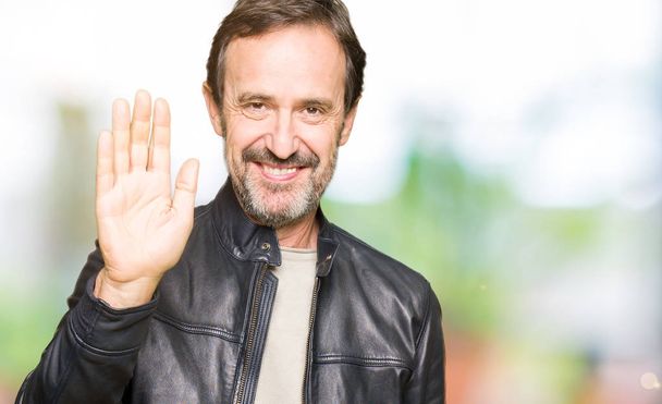 Middle age handsome man wearing black leather jacket Waiving saying hello happy and smiling, friendly welcome gesture - Photo, Image