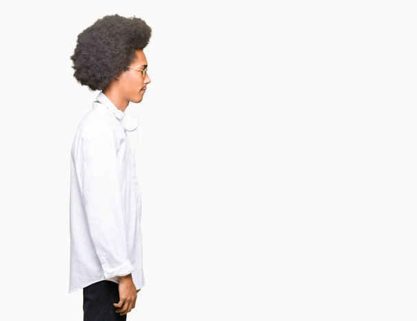 Jonge Afro-Amerikaanse man met afro haar dragen zonnebril en hoofdtelefoon op zoek naar kant, ontspannen profiel pose met natuurlijke gezicht met vertrouwen glimlach. - Foto, afbeelding