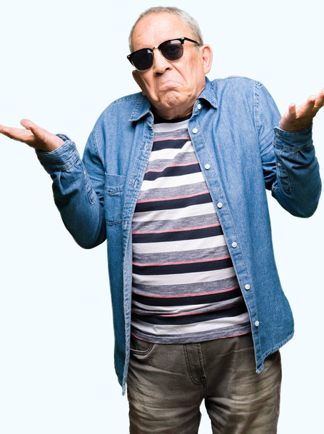 Un bel homme âgé portant une veste en denim et des lunettes de soleil s'exprime sans idée et confus avec les bras et les mains levés. Concept de doute
. - Photo, image