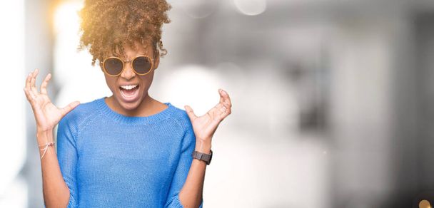 Belle jeune femme afro-américaine portant des lunettes de soleil sur fond isolé fou et fou crier et crier avec une expression agressive et les bras levés. Concept de frustration
. - Photo, image
