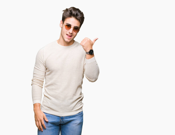 junger gutaussehender Mann mit Sonnenbrille über isoliertem Hintergrund lächelt mit glücklichem Gesicht und zeigt mit erhobenem Daumen zur Seite. - Foto, Bild