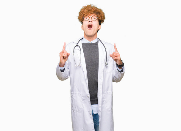 Молодой красивый врач в медицинском пальто удивлен и удивлен, глядя вверх и указывая пальцами и подняв руки
. - Фото, изображение