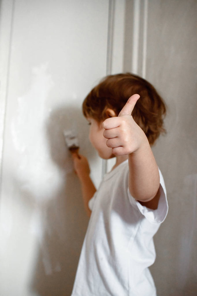 A little naughty girl in a white t-shirt paints the wall with white paint. shows class. - 写真・画像