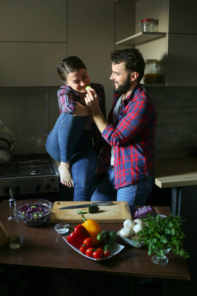 Beautiful young couple in kitchen at home while cooking healthy food. Husband cuts salad and feeds his wife cucumber. Scene from family life. - Foto, Bild