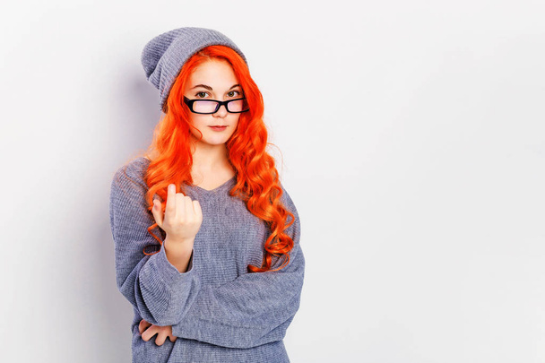Portrét dívky zrzny, která mi ukazovala gesto. Ženská modelka s červenými vlasy v pleteném svetru a s brýlemi na šedé pozadí s kopií prázdného prostoru - Fotografie, Obrázek