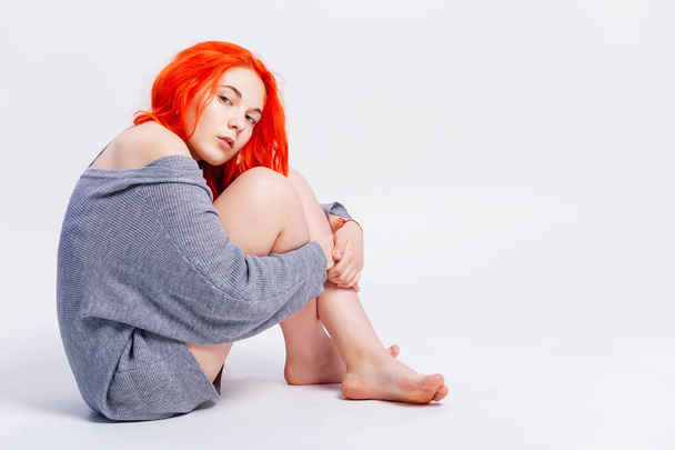 Fashion portrait of beautiful redhead teen girl dressed in knitted sweater bare legs looking over shoulder while posing on grey background sitting on floor, copy space. - Photo, Image