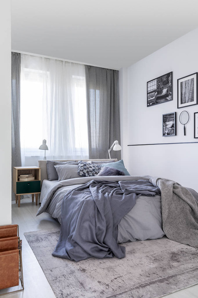 Grey sheets on bed and patterned drapes at window in bedroom interior with posters. Real photo - Fotó, kép
