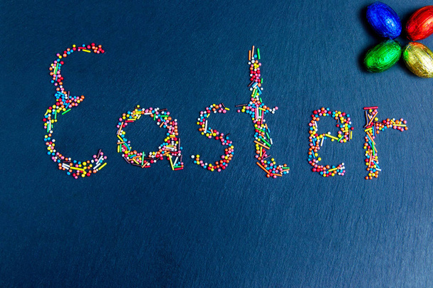 Разноцветные конфеты выкладываются в виде Пасхи на темно-синем фоне с шоколадными пасхальными яйцами
. - Фото, изображение