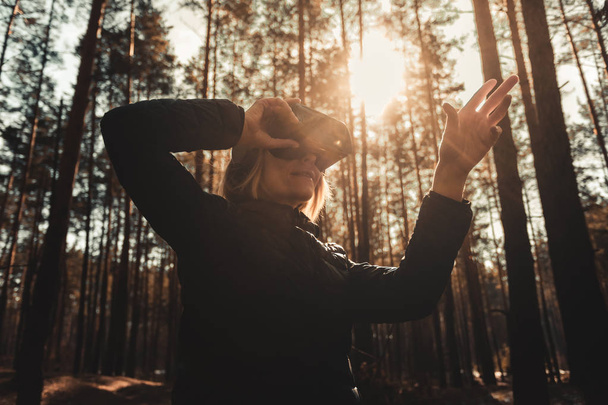 Woman in forest with virtual reality headset looking straight and trying to touch something with her hand - Photo, Image