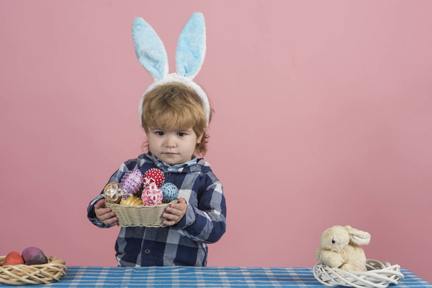 Easter present from cute kid in fluffy bunny ears. Child on pink background. Easter concept. Little boy in blue shirt holds a bowl made of straw full of Easter eggs on pink background - Photo, Image