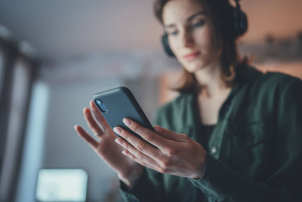 Young handsome girl relaxing at modern home,wearing casual clothes,playing music on smartphone and listening audio books in wireless headphones.Blurred background. - Фото, зображення