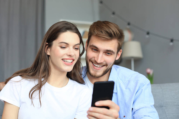 Young couple watching online content in a smart phone sitting on a sofa at home in the living room. - Zdjęcie, obraz