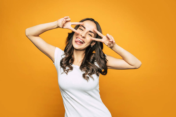 Find peace. Gorgeous young girl next to the orange-colored background with splendid smile all over her face and peace signs which she shows with her hands. - Foto, imagen