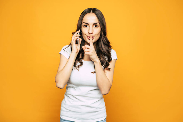 SHHH! Nice looking girl with glowing skin, slightly curled hair and elegant smile next to the flame-colored background is showing shh because she is having a phone call. - Φωτογραφία, εικόνα